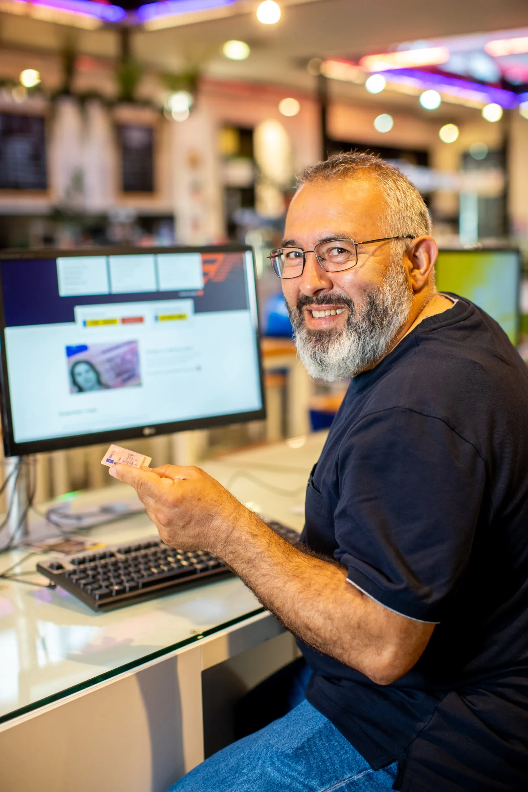 Man van ongeveer 50 jaar oud zit achter computer en heeft zijn rijbewijs in zijn linkerhand. De man kijkt lachend achterom de camera in.